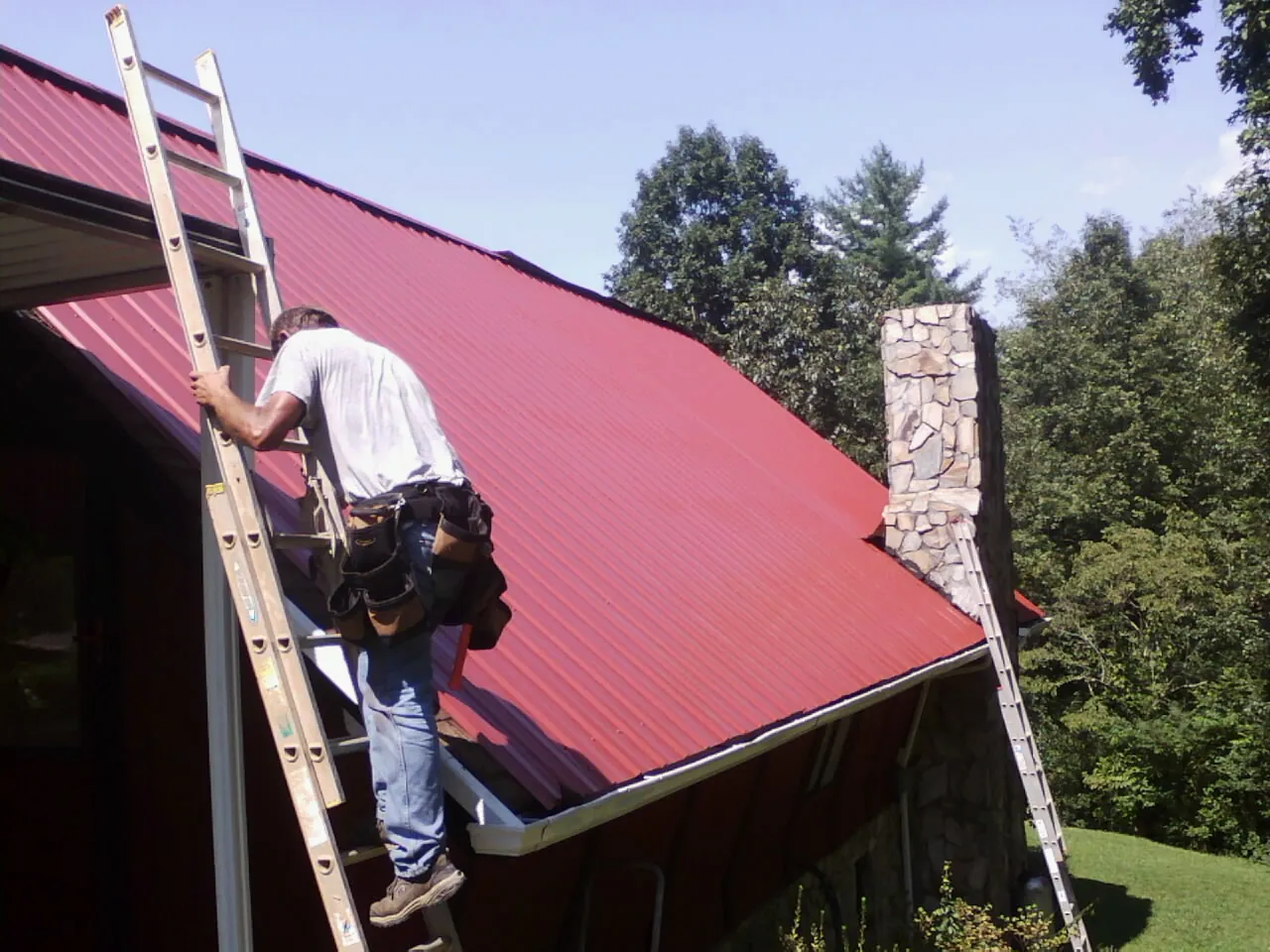 Roofer on a ladder beside a red metal roof and stone chimney — Soffit & Fascia Repair by Roofing Brothers in North Providence